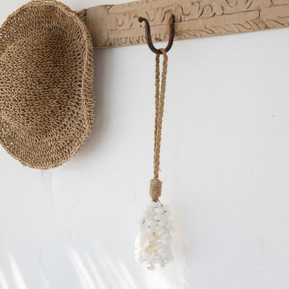 A woven straw hat hangs on a carved wooden wall hook, with a rope-and-shell hanging ornament also visible against a white wall.