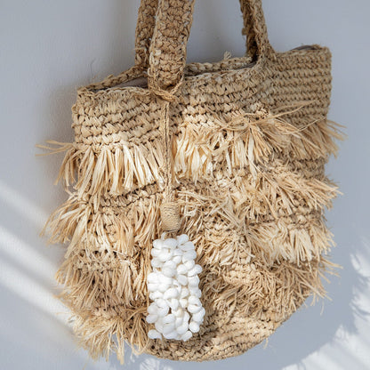A natural woven straw tote bag with layered fringe details and a white pebble bead tassel, hanging against a light gray wall with soft diagonal shadows.