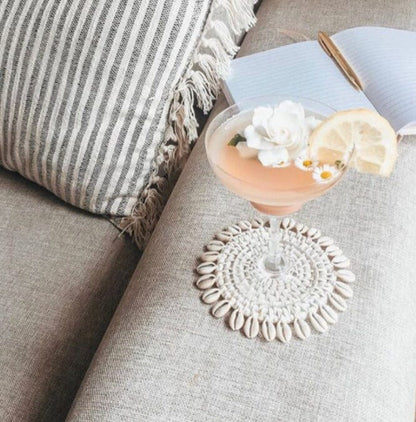 A round, white washed rattan coaster with a cowrie shell border, displayed on a sofa next to a cup with a lemon slice and a straw.