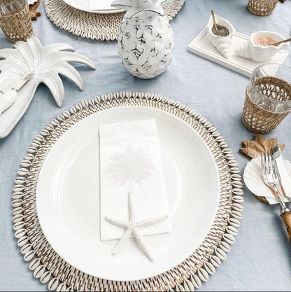 A round placemat made of natural rattan with a whitewashed finish and cowrie shell border, displayed on a table with other matching coastal-themed tableware.