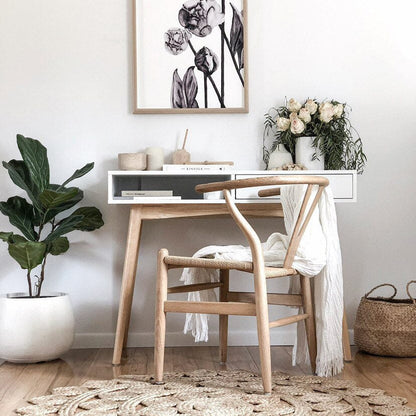 A Wishbone Replica Dining Chair in Coastal Oak finish with a natural paper cord seat and a Y-shaped backrest, placed at a dining table in a room with a white wall, green plant, and decorative items.